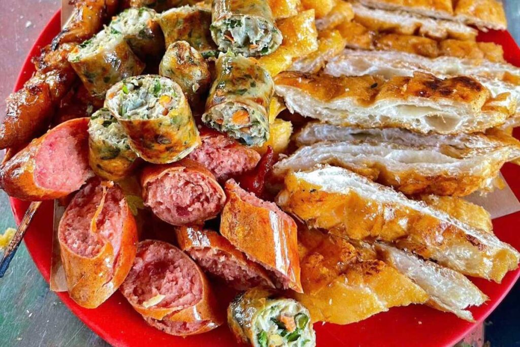Một mâm đồ ăn nướng xèo xèo vàng ruộm với các lát nem cuốn chiên, dồi sụn/xúc xích và bánh mì nướng mật ong giòn rụm được bày trên đĩa đỏ.