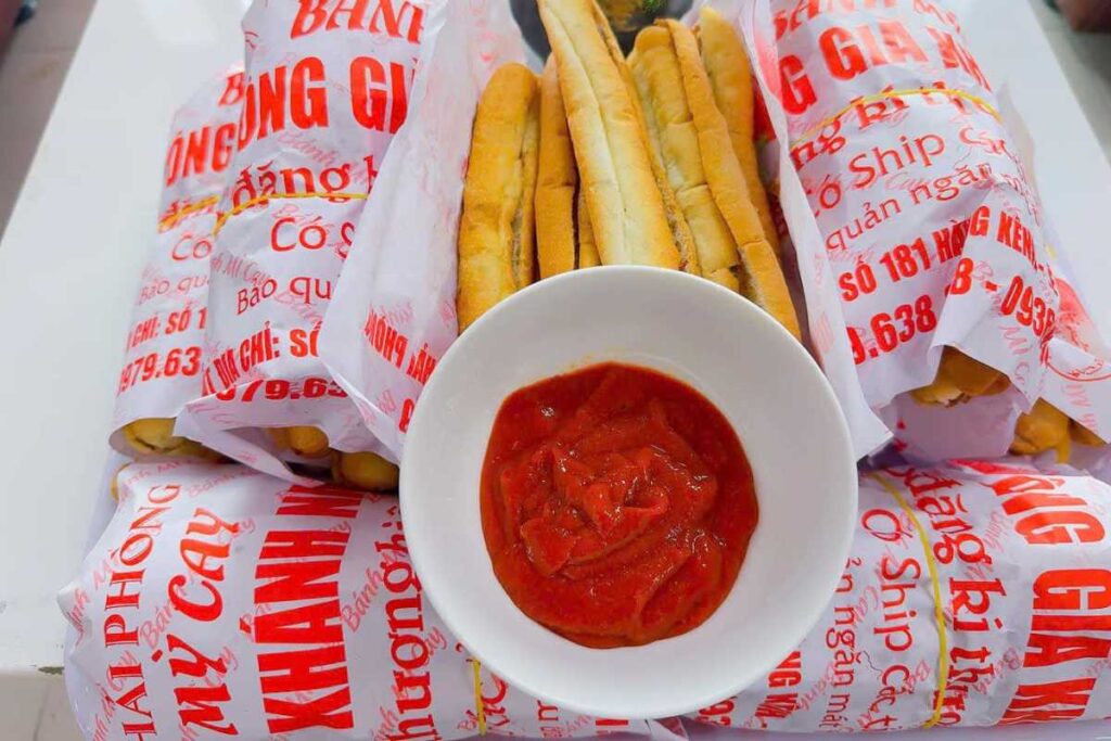 Khay bánh mì que vàng ruộm vừa ra lò bên cạnh bát tương ớt chí chương đỏ tươi tại quán Ông Già Khánh Nạp.