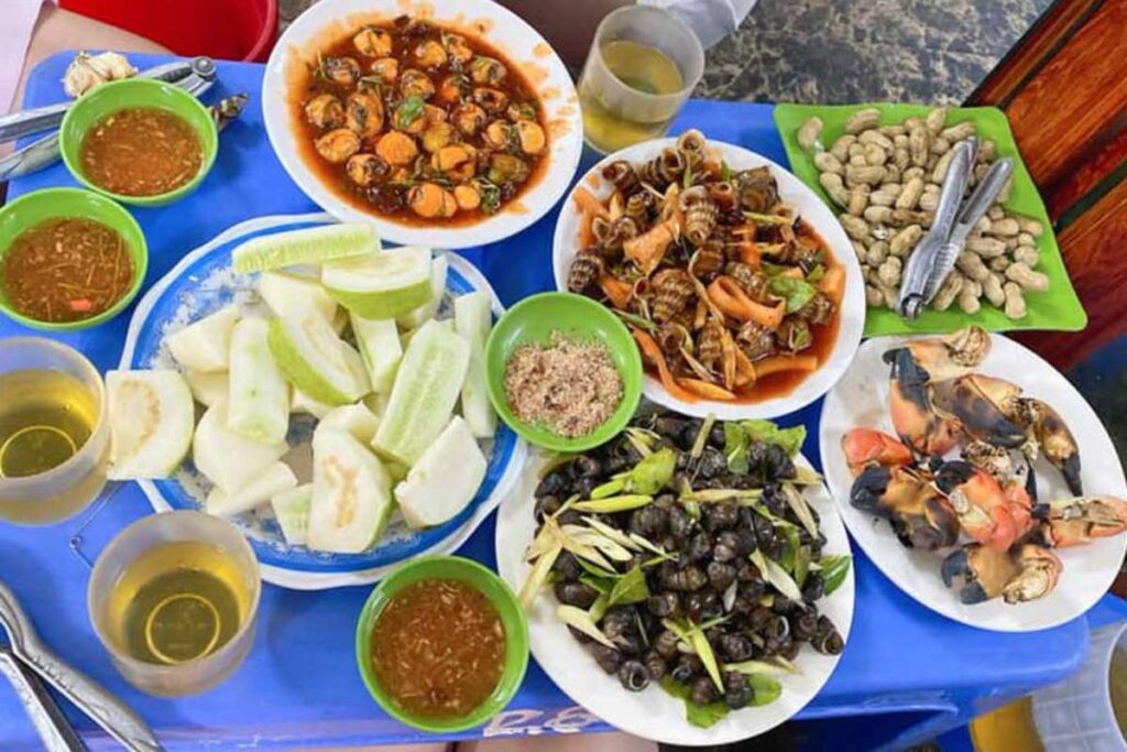 Mâm ốc thập cẩm với nhiều loại ốc xào măng cay, hàu nướng mỡ hành trên đĩa trên bàn xanh.